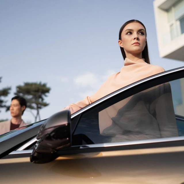 Elegant woman in a peach turtleneck stands by an open car door as a man stands in the background near a modern building.