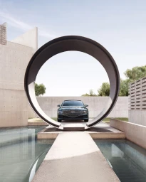A dark green luxury car parked inside a large metal ring structure with concrete architecture and water in the foreground.
