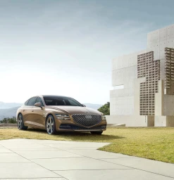 Golden luxury sedan parked on a paved forecourt beside a modern concrete building and grassy lawn under a pale sky.