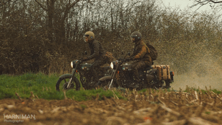 Two riders in helmets driving vintage motorcycles along a dirt path beside a field of sparse trees.