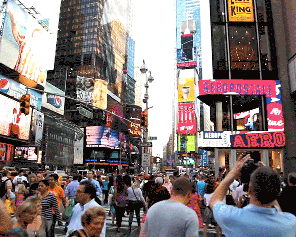Crowd of people walks through Times Square in New York City, surrounded by bright billboards and skyscrapers, with traffic lights.