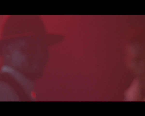 Red-toned blurred image with two partially visible figures, one in a hat, against a plain background.