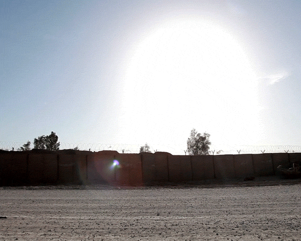Sunlit outdoor scene with a long concrete wall topped with barbed wire, set against a bright sky and a ground area with trees.