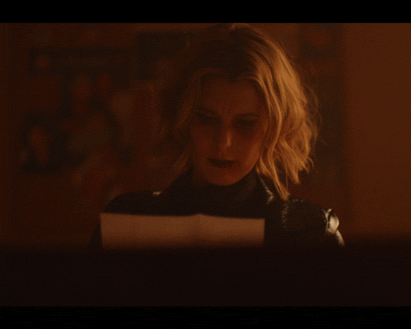 A woman with short blonde hair reads a white paper in warm lighting, wearing a black top, with a blurry background.