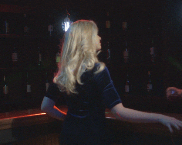 Blonde woman in a blue dress stands at a bar with bottles on a shelf in the background, illuminated by warm, dim lighting.