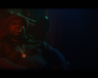 Man wearing dark shirt and hat seated in a dimly lit room with moody red and blue lighting by photographer Patrick House