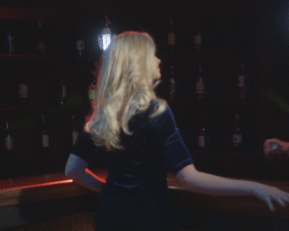 Blonde woman in a dark dress standing by a bar counter in a dimly lit venue photographed by Patrick House