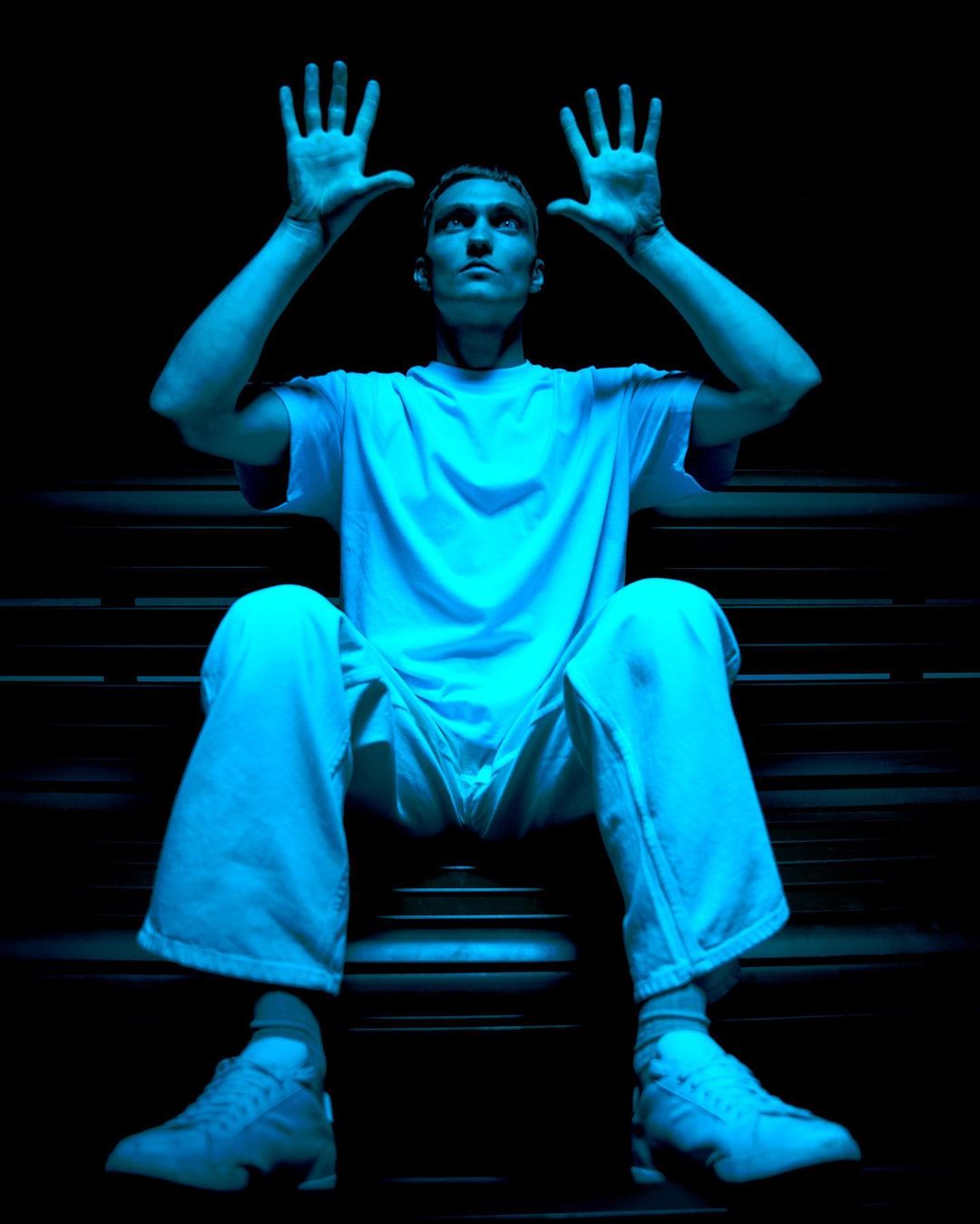 Man sits on steps with his hands up, wearing a white t-shirt, pants, and shoes, and bathed in bright blue light against a black background.