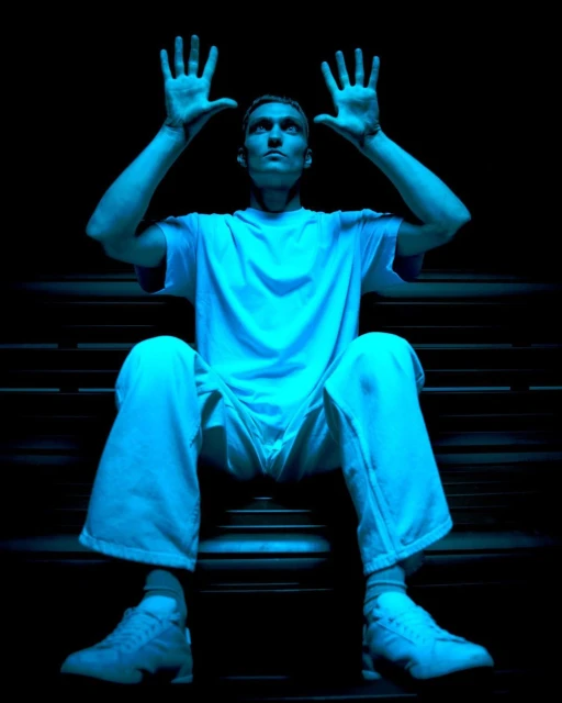 Man sits on steps with his hands up, wearing a white t-shirt, pants, and shoes, and bathed in bright blue light against a black background.