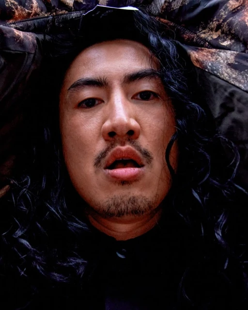 Headshot of an adult male with long black hair, a mustache, and a goatee, looking directly at the camera from a low angle in dark lighting.