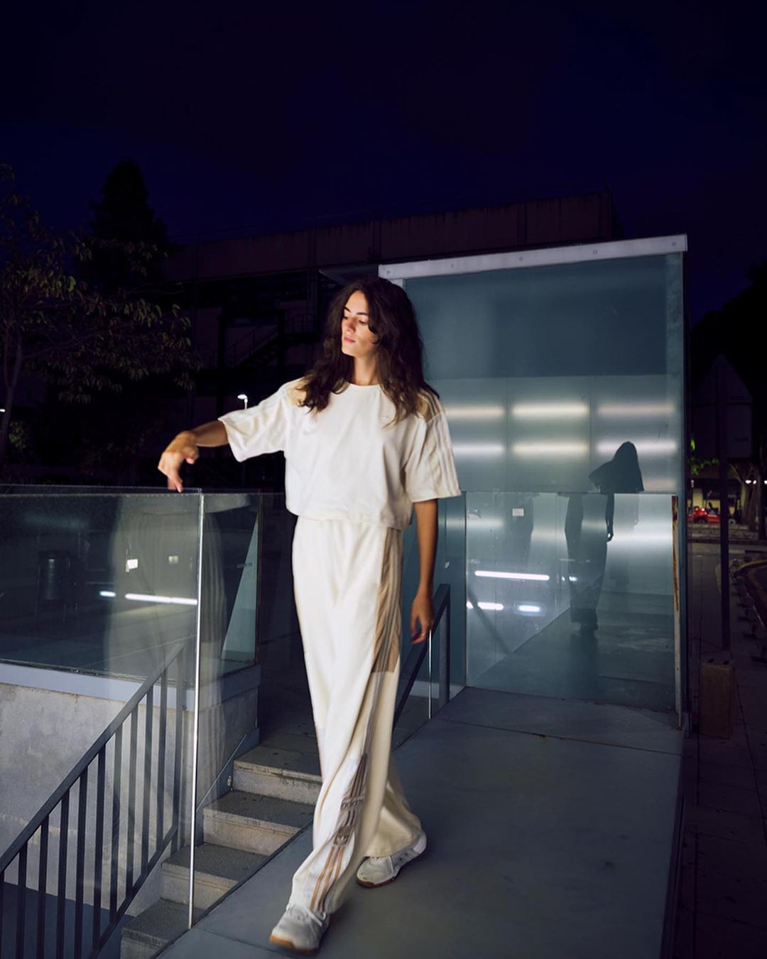 Young woman in loose white outfit poses on stairs outdoors at night, showcasing contemporary fashion against a modern architectural backdrop.