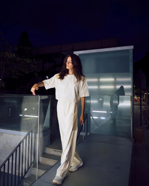 Young woman in loose white outfit poses on stairs outdoors at night, showcasing contemporary fashion against a modern architectural backdrop.