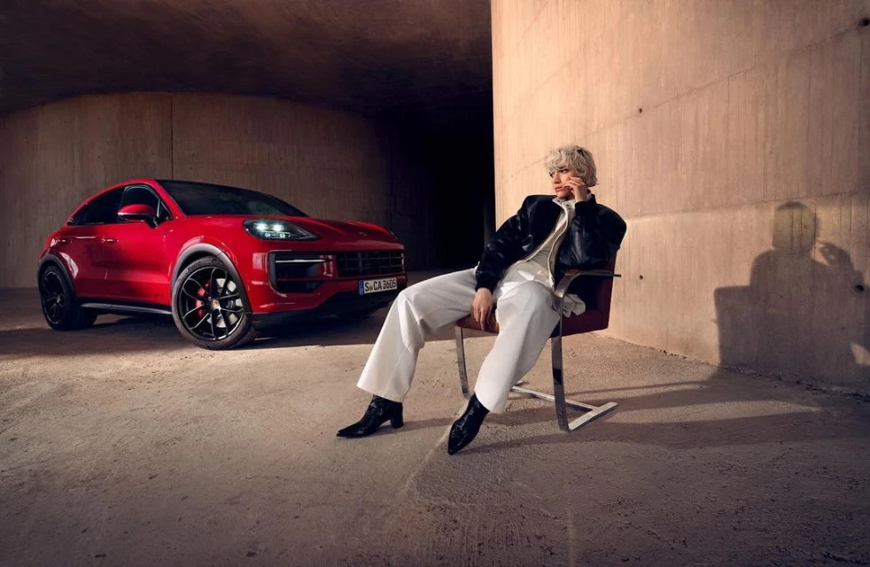 Person sits in chair next to red Porsche coupe in industrial setting with neutral concrete walls and floor in a lifestyle shot.