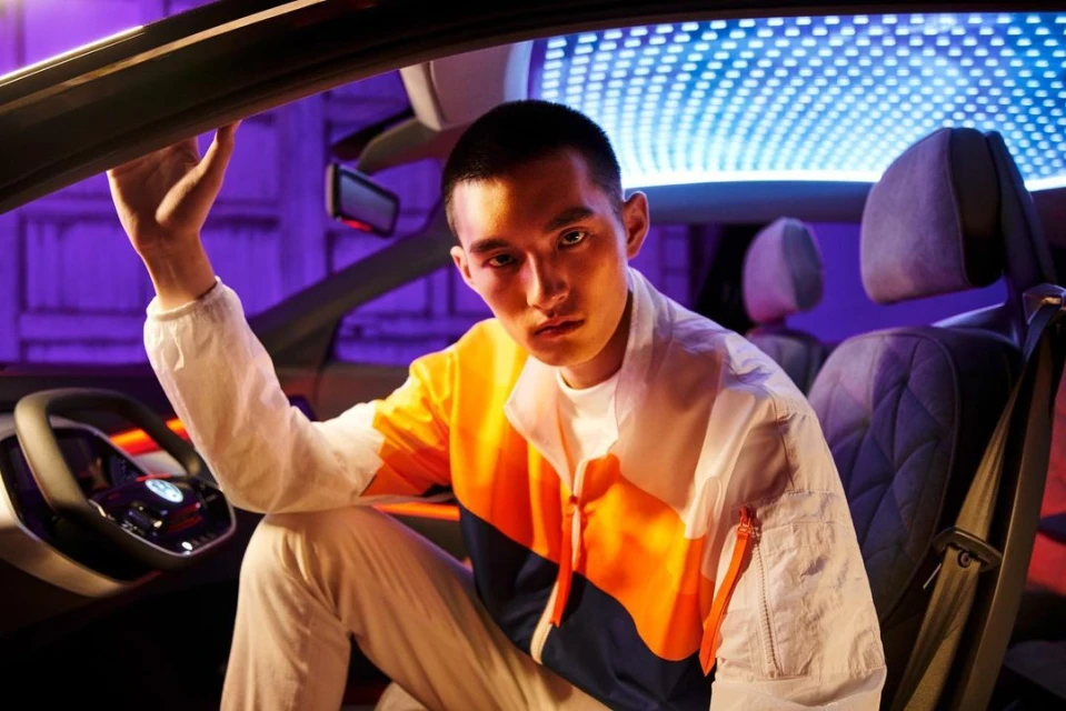 Adult male poses inside a modern car. Man wears a color-blocked jacket. Car interior features blue LED lighting. Modern design is visible.