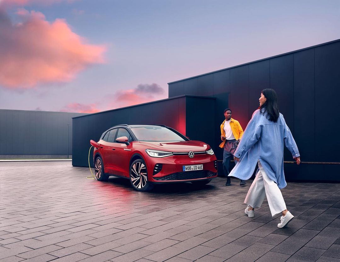 Red Volkswagen ID.4 electric car charging with two people walking on a brick surface in front of a dark building at dusk.