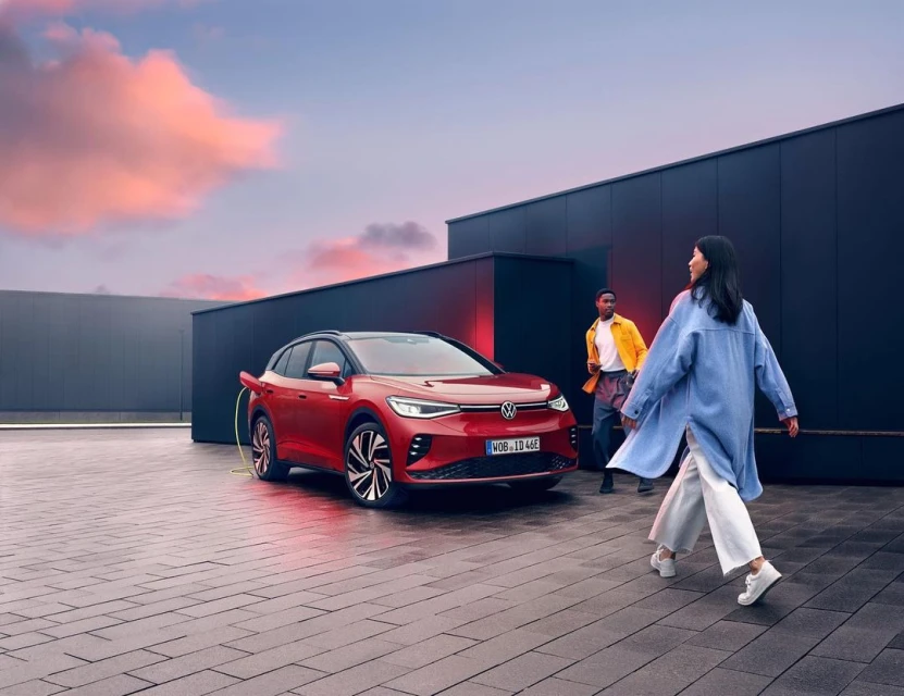 Red Volkswagen ID.4 electric car charging with two people walking on a brick surface in front of a dark building at dusk.