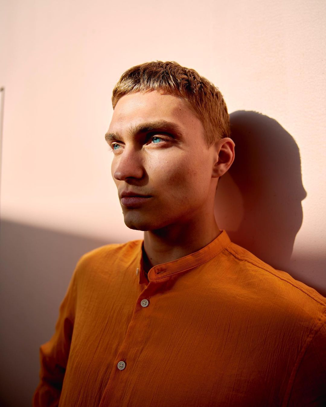 Portrait of a young adult male with short ginger hair and blue eyes wearing an orange button-down shirt against a peach-colored wall.