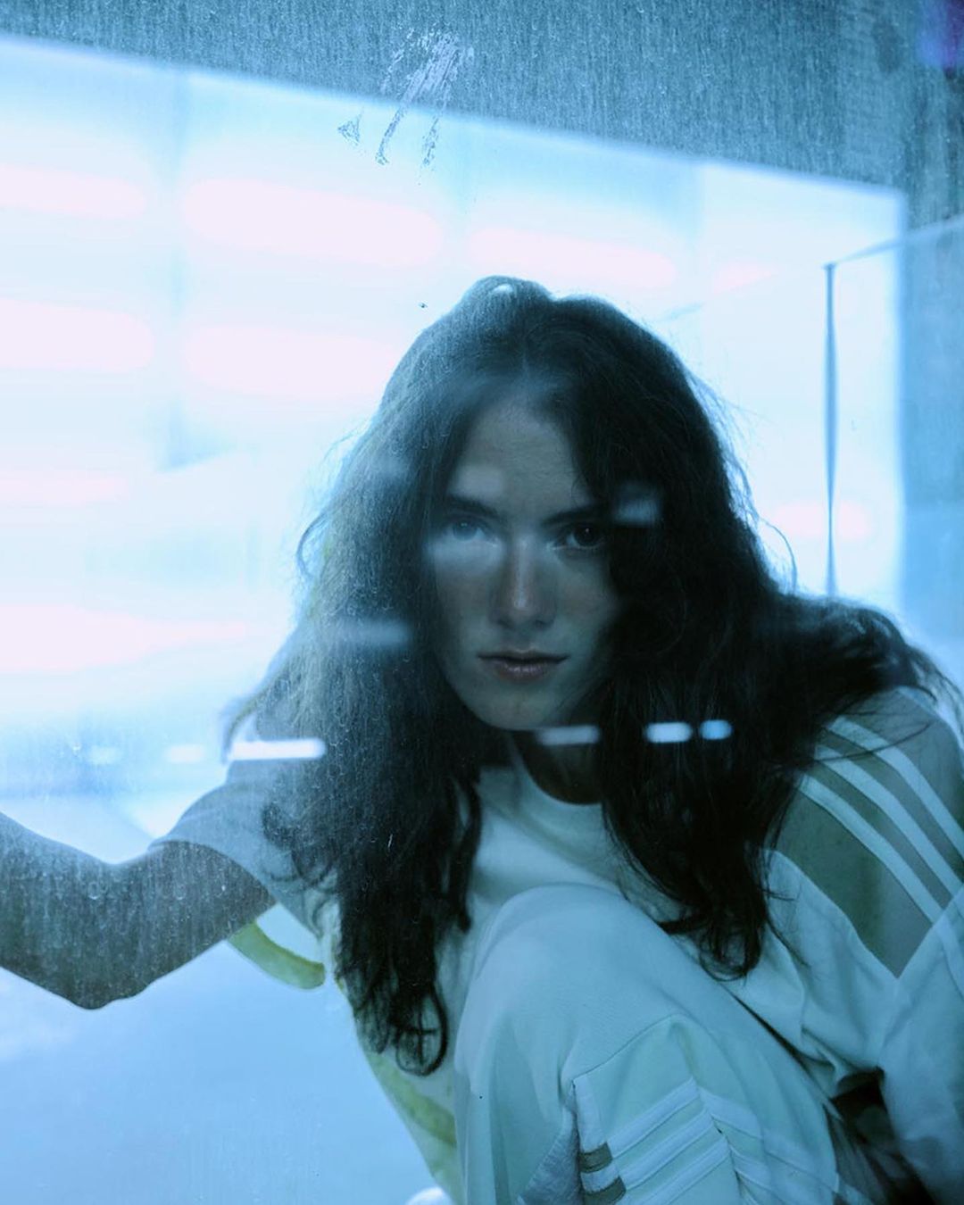 Woman with long dark hair in white athletic wear poses indoors with soft, diffused lighting, looking directly at the camera.