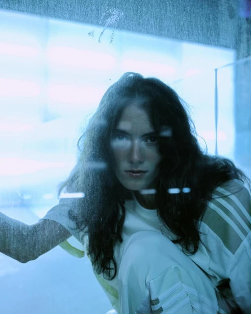 Woman with long dark hair in white athletic wear poses indoors with soft, diffused lighting, looking directly at the camera.