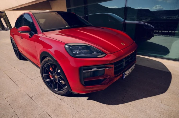 A red Porsche Cayenne GTS parked on a paved surface outside a modern building on a sunny day, reflecting another car in the window.