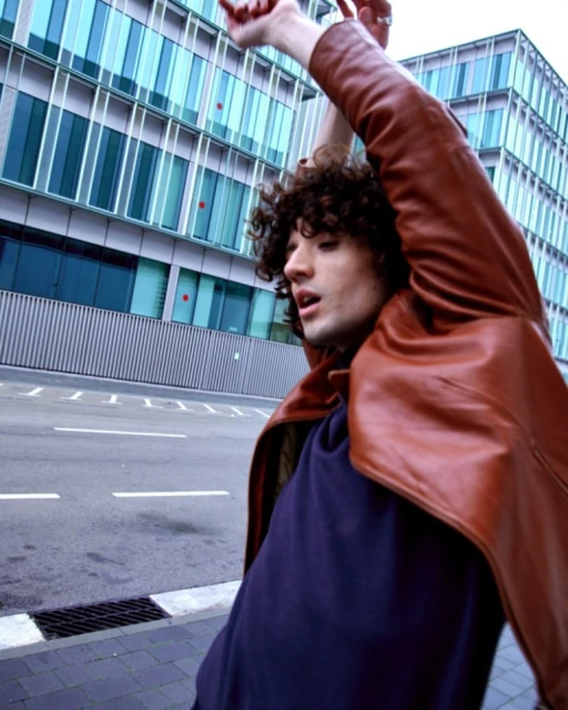 Young adult with curly hair wearing brown leather jacket and navy shirt in urban outdoor scene with modern building background.