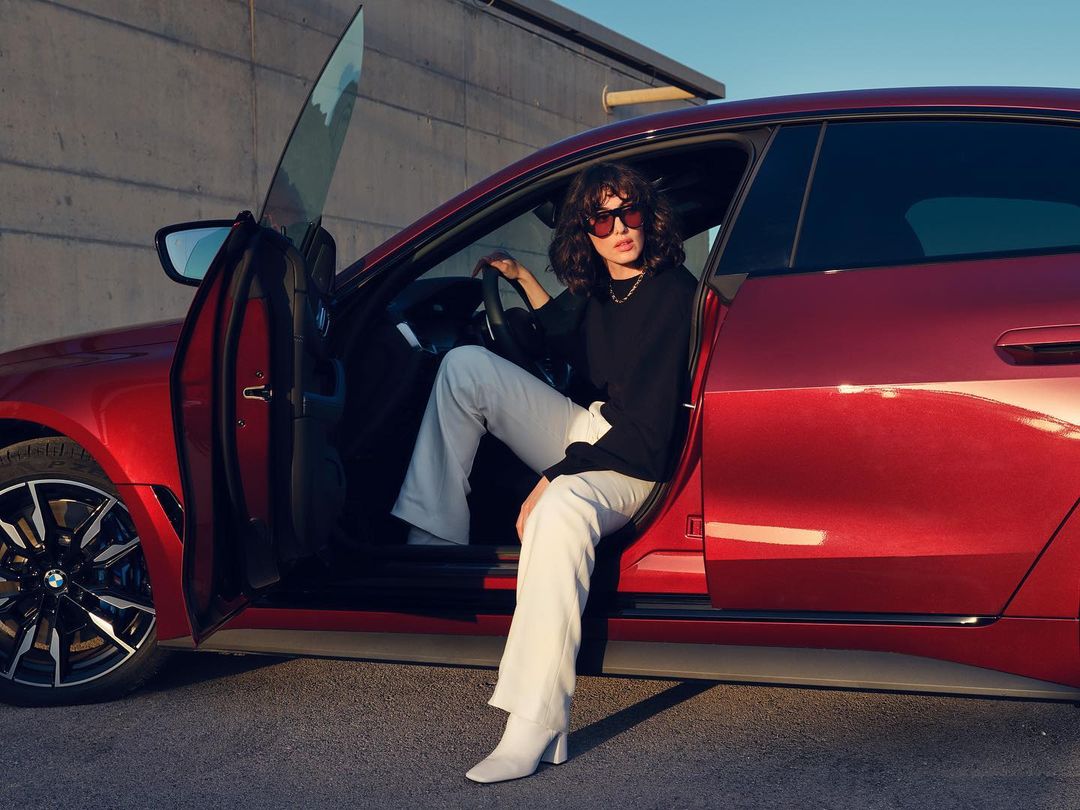 A woman in sunglasses is seated in the doorway of a red BMW car in front of a concrete wall on a sunny day.