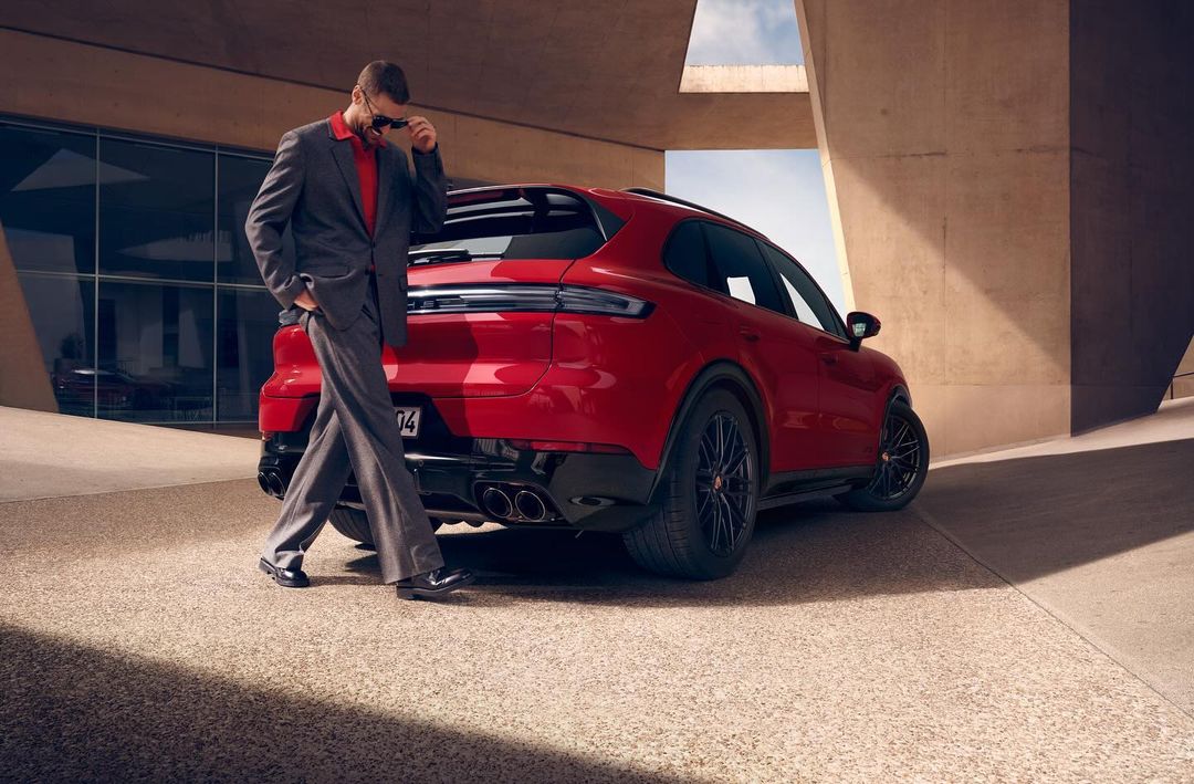 A red Porsche Cayenne parked outdoors with a well-dressed man in a suit standing next to it, holding sunglasses, against a building backdrop.