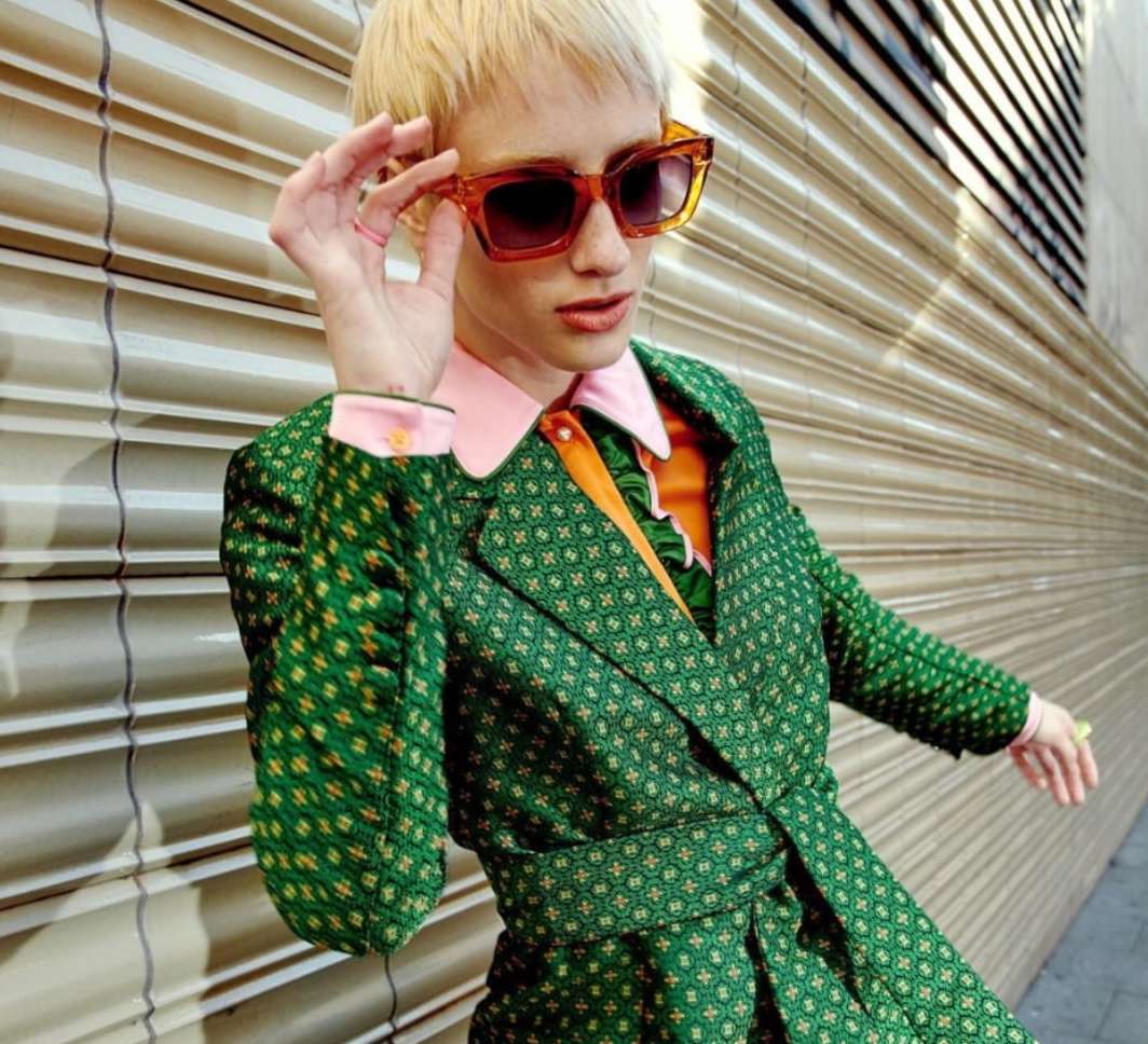 Woman wearing oversized sunglasses and a green patterned suit poses against a corrugated metal wall in a medium shot.