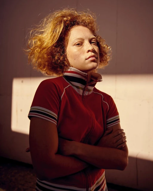 Portrait of a young adult with curly red hair, wearing a red jacket, arms crossed, against a cream-colored wall with natural light.