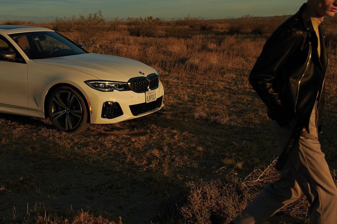 White BMW car parked in arid landscape next to a person in black leather jacket and tan pants walking across dry grass.