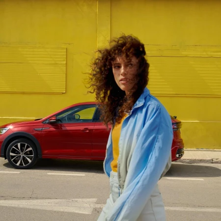 Curly-haired woman in a light blue jacket walking past a red car parked against a bright yellow wall