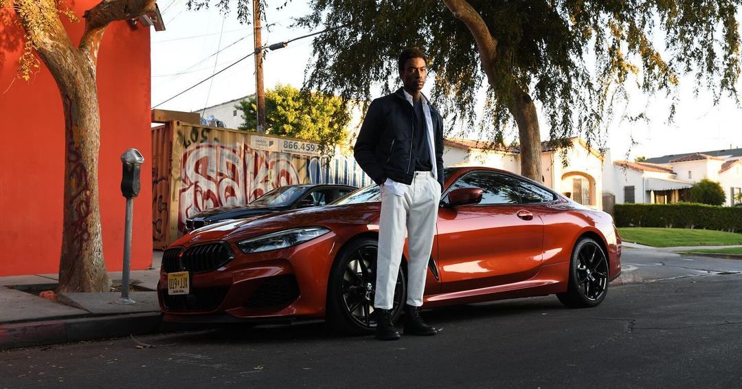 A fashionable African-American man poses near an orange BMW on a tree-lined street with houses in the background on a sunny day.