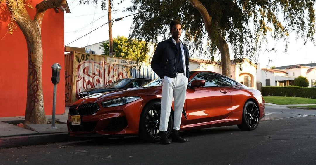 A fashionable African-American man poses near an orange BMW on a tree-lined street with houses in the background on a sunny day.