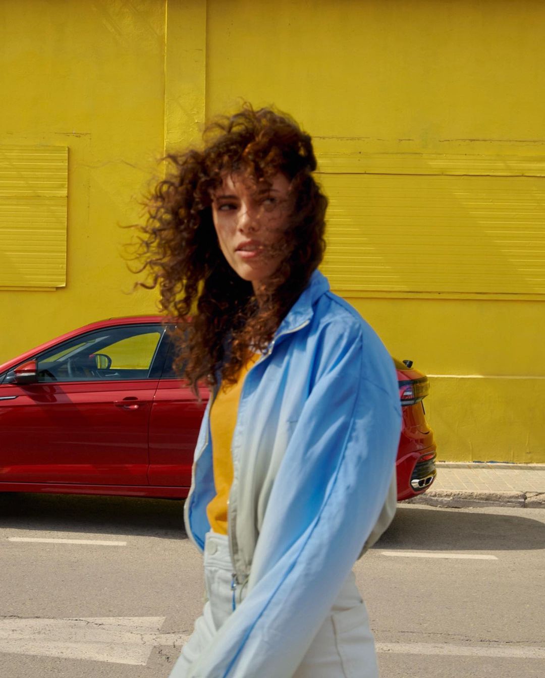 Young woman with curly hair poses on a city street next to a red car with a yellow building wall in the background on a sunny day.