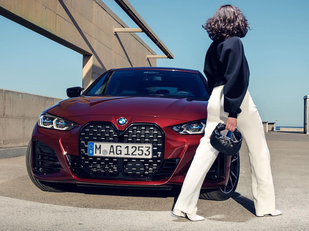 Burgundy BMW i4 electric vehicle parked on a concrete surface with a woman in a black sweater and white pants holding a purse.