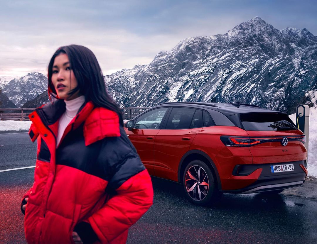 Asian woman in red puffer jacket poses with a red Volkswagen ID.4 against a snowy mountain backdrop on an overcast day.