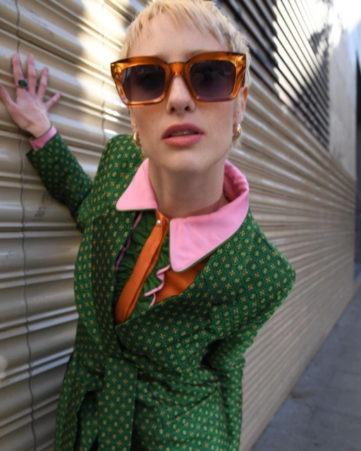 Woman in green patterned jacket and orange sunglasses poses against gray corrugated wall.  She is adorned with gold hoop earrings and rings.