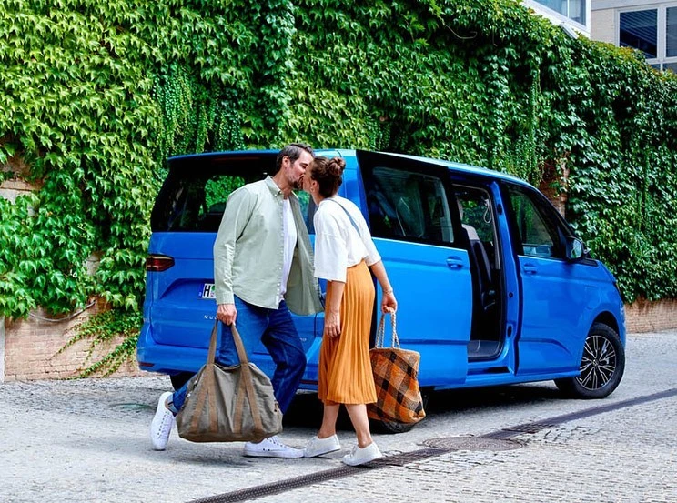 A couple kisses next to a bright blue van, standing on a stone path, holding tote bags with a green ivy-covered wall in the background.
