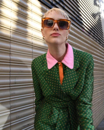 Blonde woman wearing amber sunglasses and a green patterned blazer with a pink collar standing by a metal shutter.