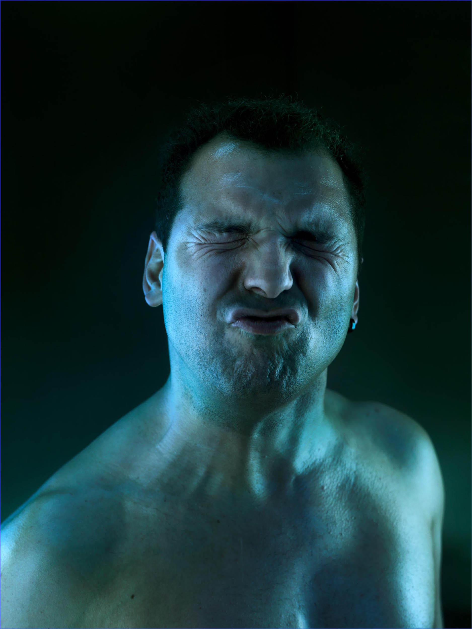 Adult male grimaces with eyes closed in a studio portrait with green and blue lighting effects against a dark backdrop, showing intense emotion.