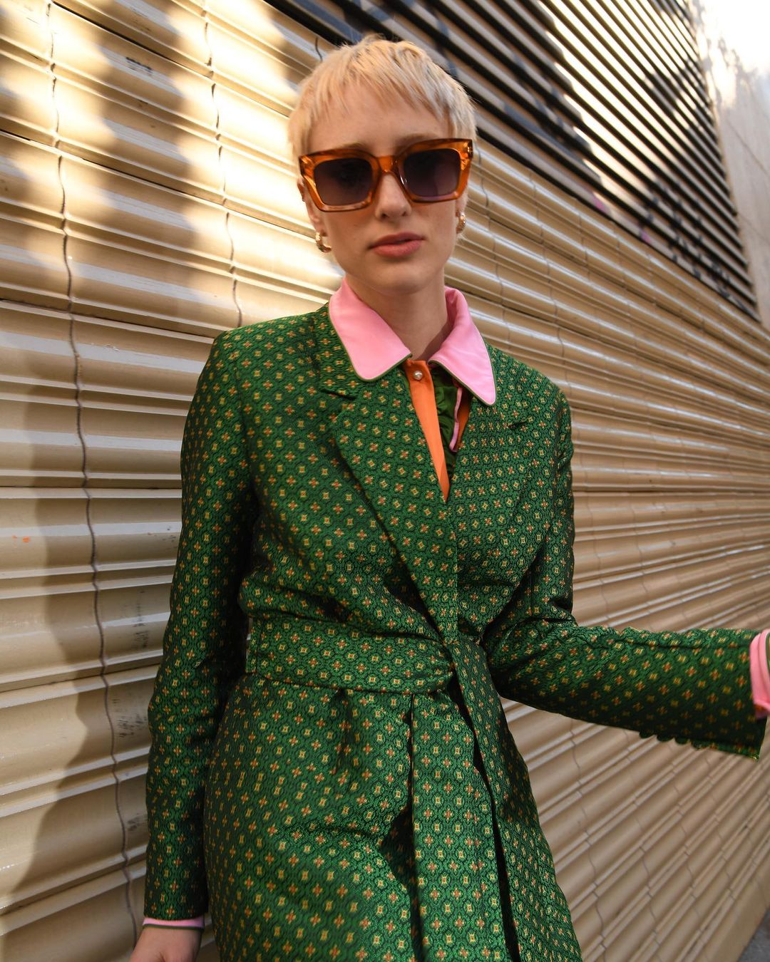 Woman in green patterned blazer and orange sunglasses standing outdoors in front of a closed metal shutter door.
