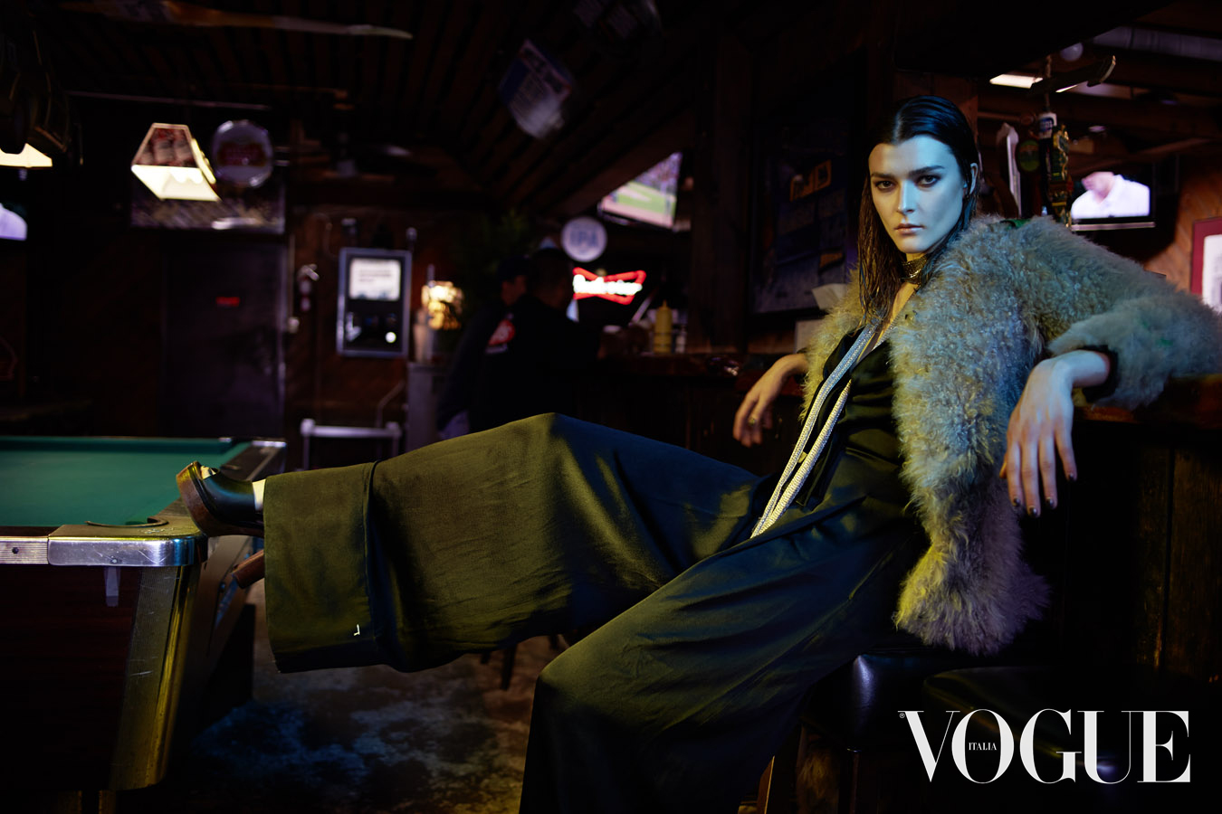 Fashion editorial image featuring a woman in a jacket and wide-leg pants, seated in a bar with a pool table, Vogue Italia logo visible.