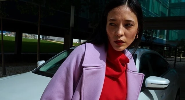 Woman in a lavender coat poses with a white car in an urban setting, showcasing fashion and automotive elements.