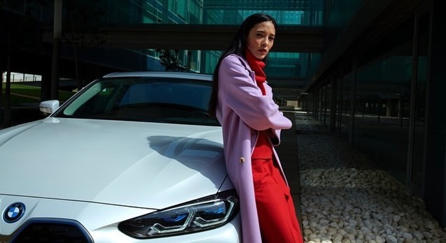 Woman in coat poses next to a white BMW car in front of a modern office building on a sunny day in a medium outdoor shot.
