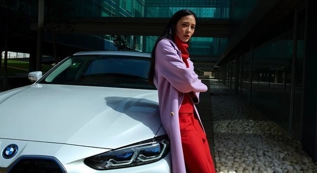 Woman in coat poses next to a white BMW car in front of a modern office building on a sunny day in a medium outdoor shot.