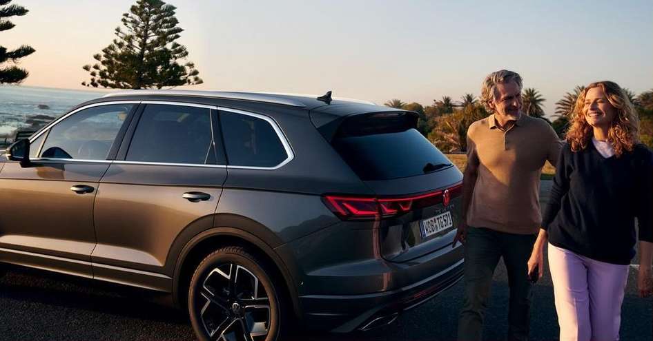 Grey SUV with an adult couple standing near it on a road near the ocean with trees and a sunset in the background.