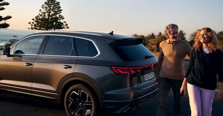 Grey SUV with an adult couple standing near it on a road near the ocean with trees and a sunset in the background.