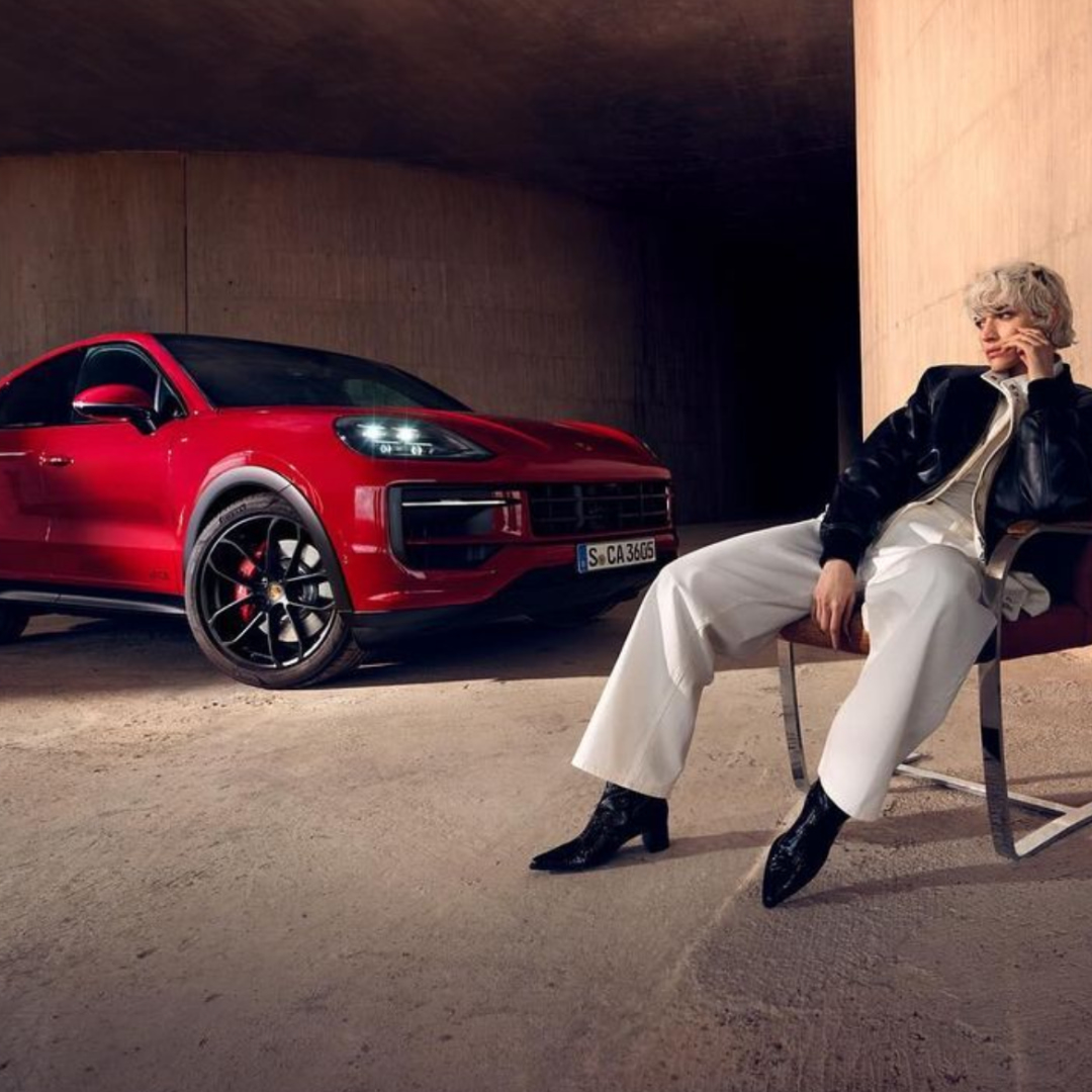 A red Porsche SUV sits next to a fashionably dressed woman sitting on a metal chair in an indoor setting with soft lighting.