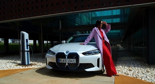 Woman in red dress and lilac coat poses with a white BMW i4 electric car in front of a modern building on a sunny day.