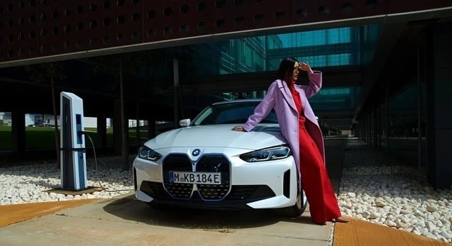 Woman in red dress and lilac coat poses with a white BMW i4 electric car in front of a modern building on a sunny day.